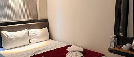 A hotel room with a bed, a red throw blanket, a white towel, a bedside table with a water bottle, a kettle, and a cup.