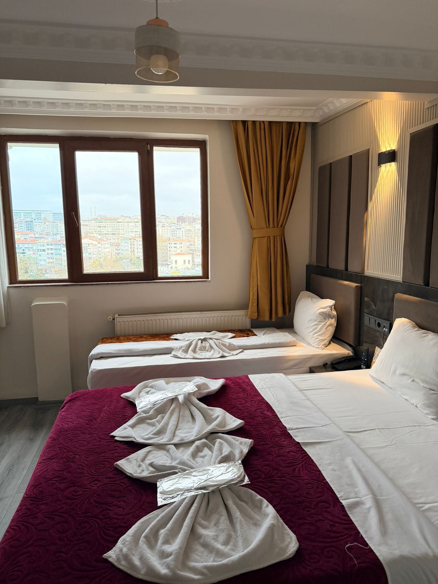A hotel room with two beds, a large window with a city view, a radiator, and a hanging light fixture.