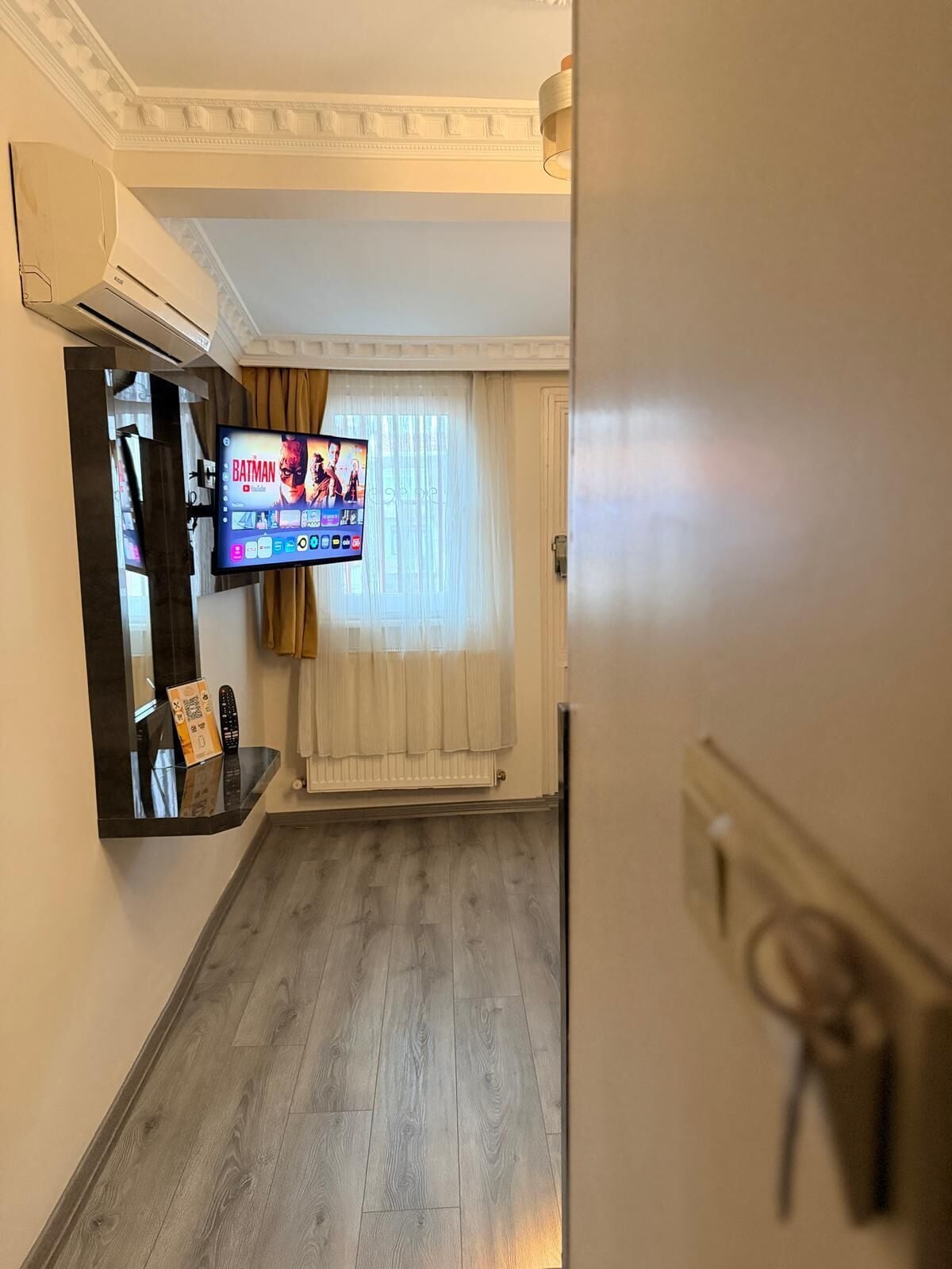 Hotellihuone, jossa on seinälle asennettu taulutelevisio, puulattia ja ikkuna, jossa on ohuet verhot.