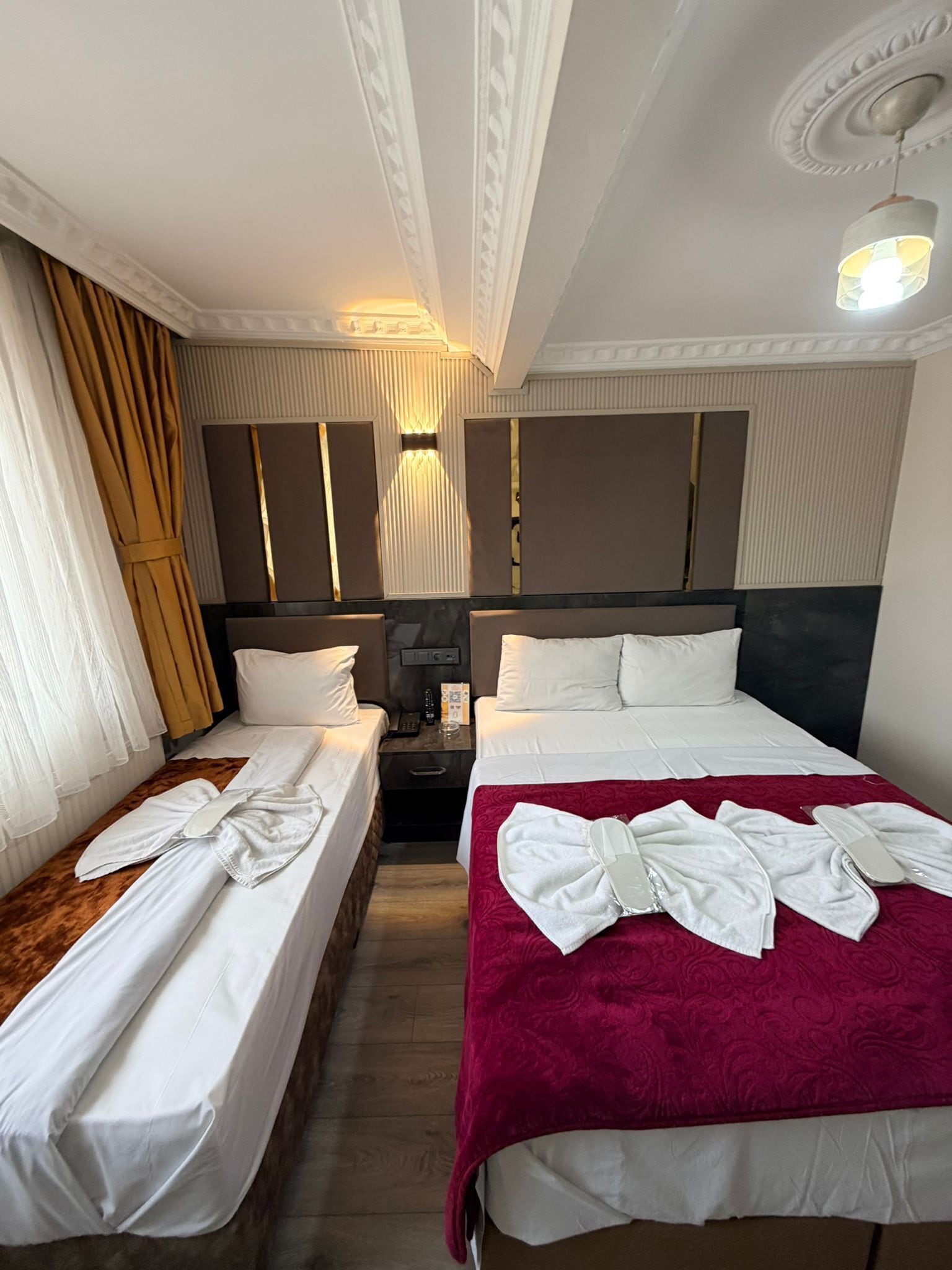 A hotel room with two beds, a red bedspread, white linens, a wooden nightstand, a wall-mounted lamp, and a ceiling with decorative molding.