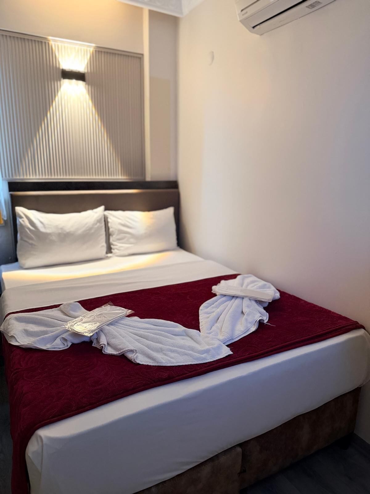 A hotel room with a bed, pillows, a red blanket, and a wall-mounted air conditioner.