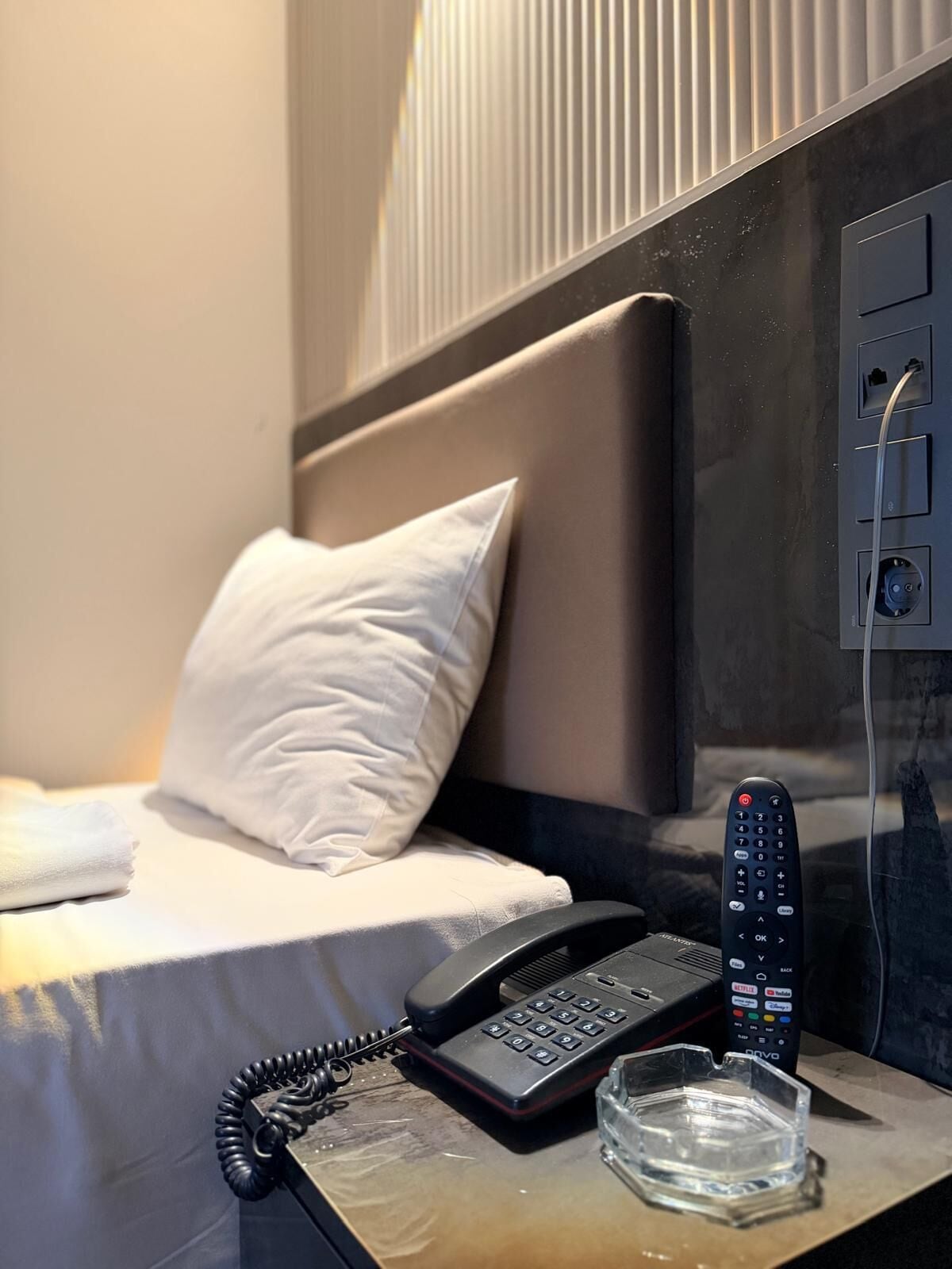 A hotel room with a bed, a bedside phone, a remote control, and a glass of water on a nightstand.