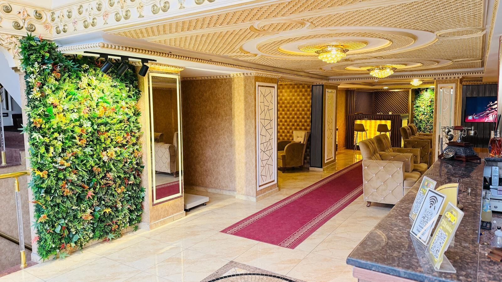 En hotelllobby med en röd matta, en receptionsdisk, en sittgrupp och en vägg som är dekorerad med grönska.