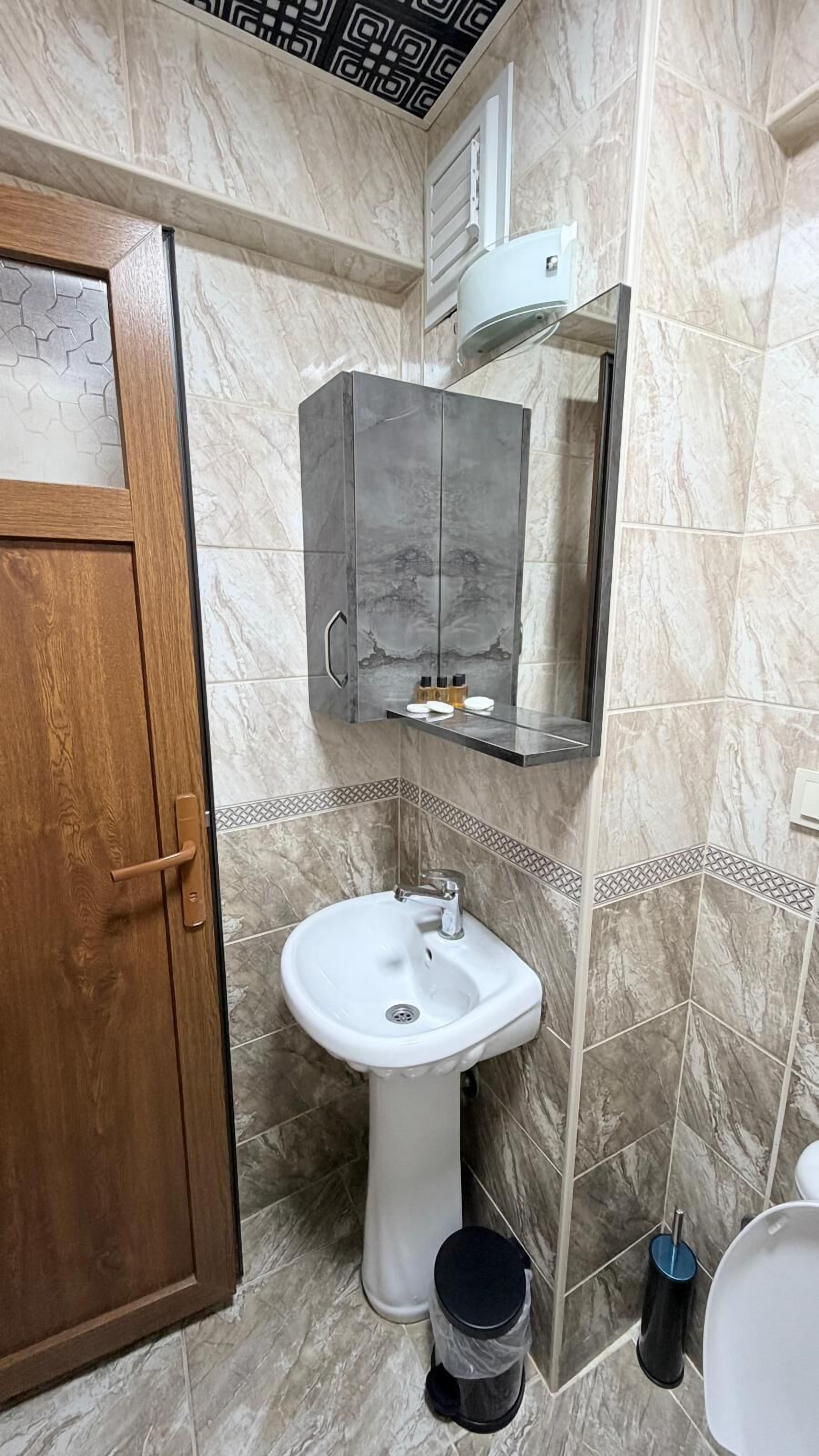 A bathroom with a wooden door, a white pedestal sink, a mirror above the sink, and a toilet.