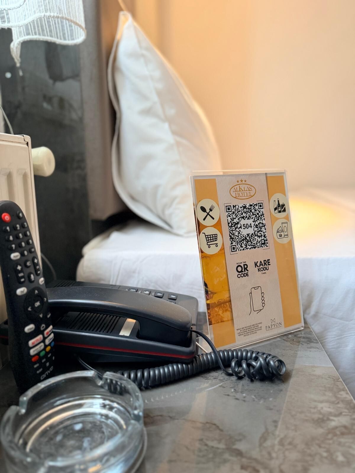 Hotelkamer met een bed, nachtkastje met telefoon, glazen asbak en een QR-code bord.