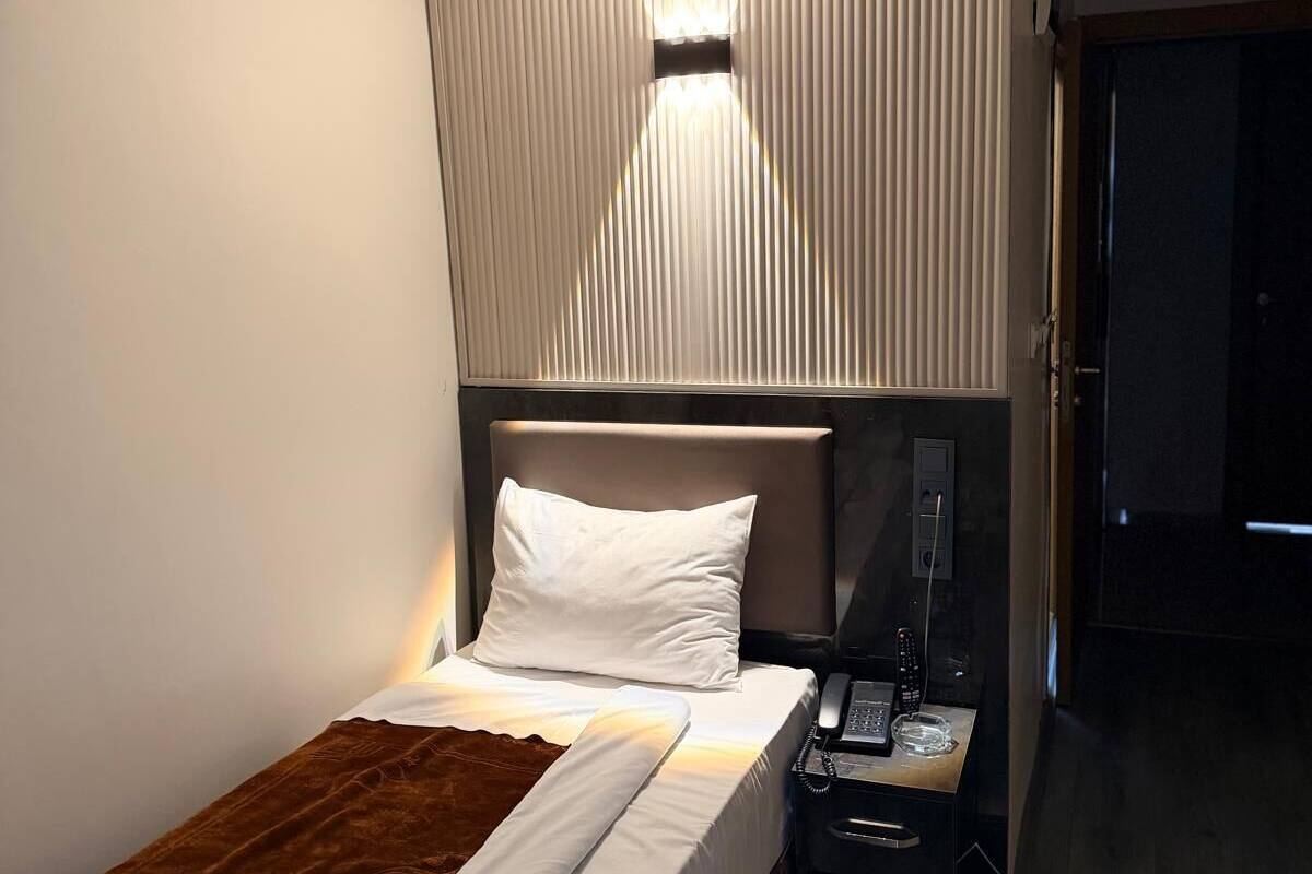 A neatly made bed with a brown throw, a bedside table with a phone and remote, and a wall-mounted light fixture.