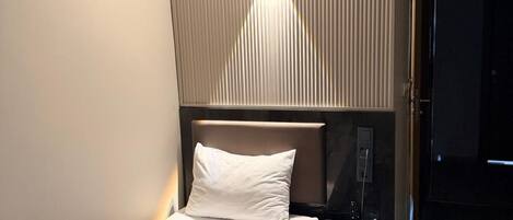 Un letto rifatto con cura, una coperta marrone, un comodino con telefono e telecomando, e un punto luce fissato al muro.