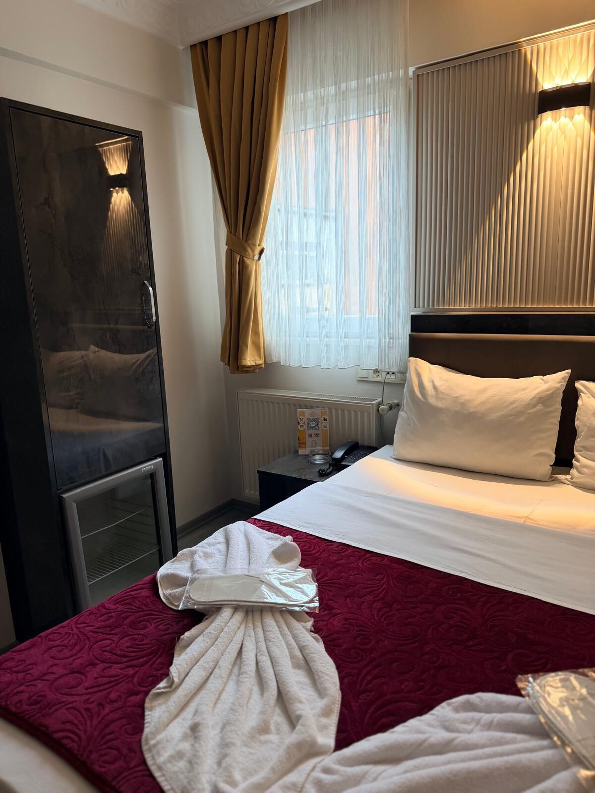 A hotel room with a bed, a wardrobe, a radiator, a bedside table with a phone, and a window with curtains.