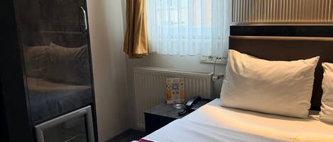 A hotel room with a bed, a wardrobe, a radiator, a bedside table with a phone, and a window with curtains.