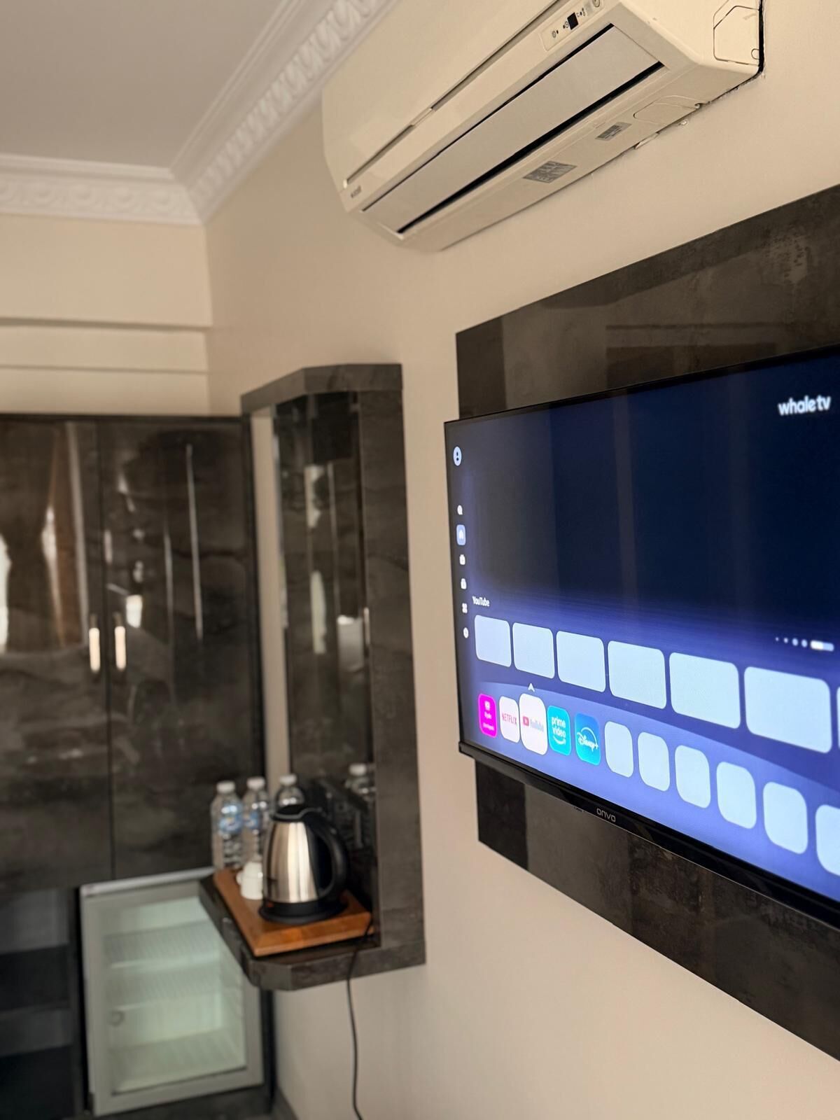A modern hotel room with a mounted flat-screen TV displaying a YouTube interface, a built-in air conditioning unit, and a small refrigerator.