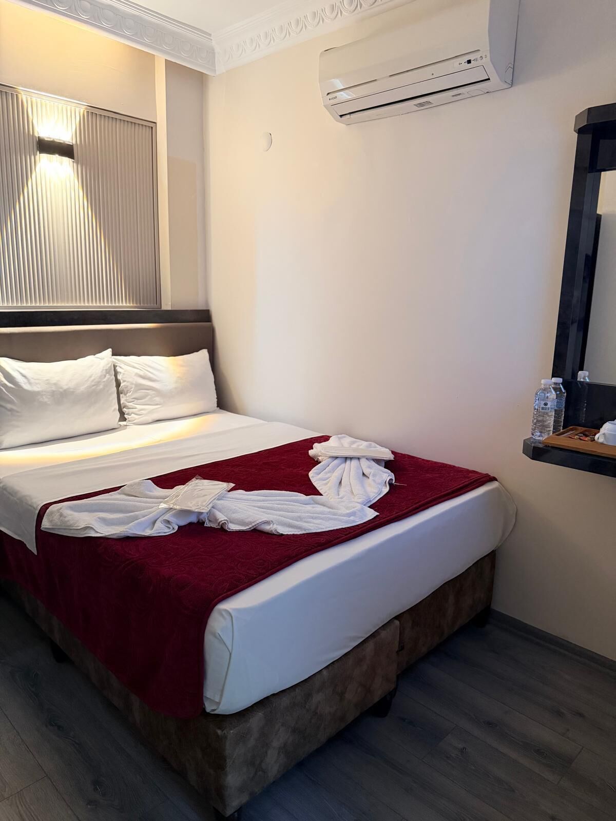 A hotel room with a bed, white pillows, a red blanket, a water bottle, and an air conditioning unit.