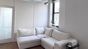 A modern living room with a white sectional sofa, a small black side table, and a window with a view of buildings. - Charming 2-bedroom Tribeca apartment with views (New York)