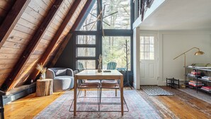 Acogedor dormitorio en ático con techo de madera, una mesa de comedor con sillas, una estantería y un amplio ventanal con vistas a árboles.