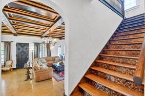 Una sala de estar con un sofá, una mesa de centro y una araña. Se observa una escalera con detalles de madera decorativos.