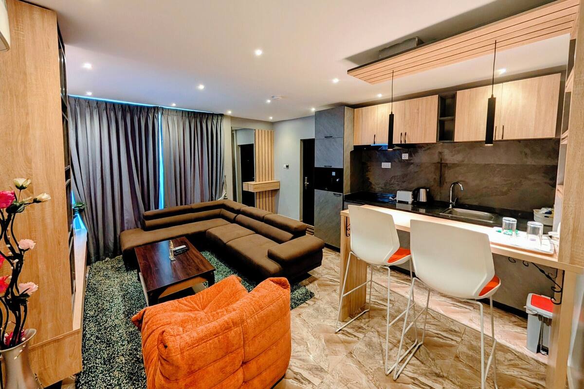 Un salon moderne avec un canapé d’angle, un petit fauteuil orange et un coin repas, la cuisine étant visible au fond.