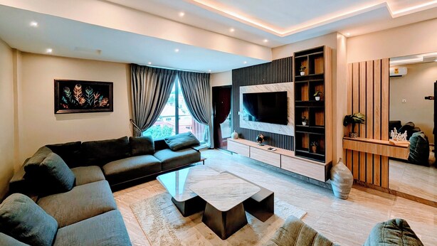 A modern living room with a sectional sofa, a flat-screen TV, a coffee table, and a built-in entertainment unit. - Condos De La ChiChi (Kumasi)