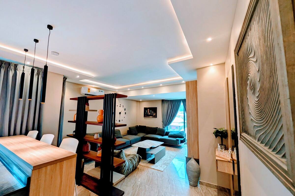 Un salon moderne avec un canapé, une table basse et un téléviseur. Il y a également un coin repas avec une table et des chaises. La pièce est dotée d'une grande fenêtre et d'un élément décoratif mural.