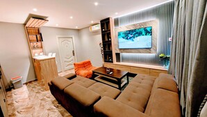A modern living room with a sectional sofa, a flat-screen TV mounted on the wall, and a built-in bookshelf. - Condos De La ChiChi (Kumasi)