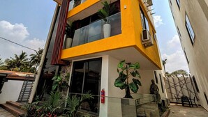 A modern multi-story building with a yellow facade, large windows, and a balcony with potted plants. - Condos De La ChiChi (Kumasi)