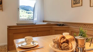 A dining area with a wooden bench, a table with a white cup and saucer, bread, a sausage, and a metal pot.