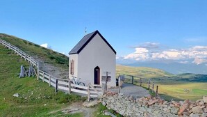 A small white church with a steep roof, located on a grassy hill with a stone wall in the foreground and a scenic view of mountains in the background.