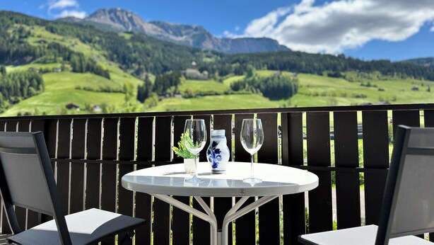 A balcony with a table set for two, featuring a vase and wine glasses, overlooking a mountainous landscape.