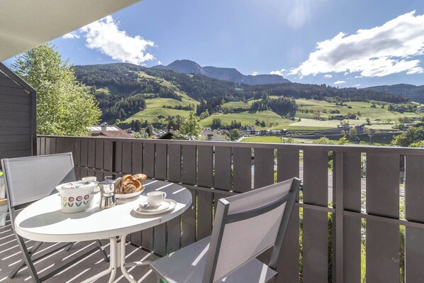 Ein Balkon mit Tisch für zwei Personen, mit Blick auf eine malerische Landschaft aus grünen Hügeln und einem kleinen Dorf.