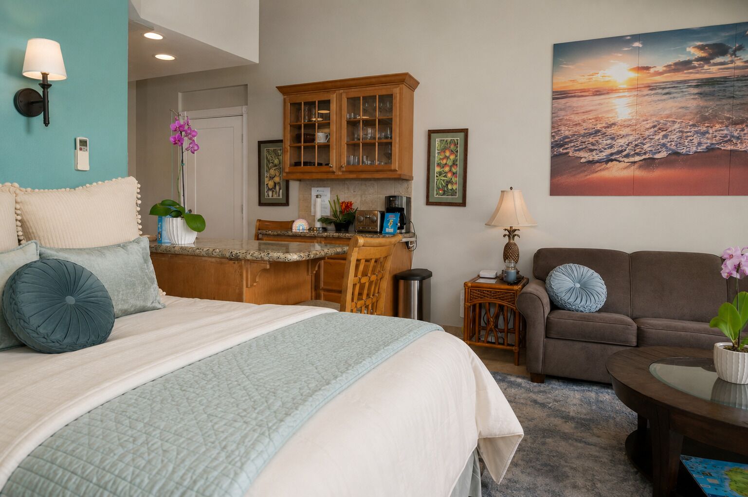 Une chambre avec un lit, un coin salon avec un canapé, un coin repas avec un comptoir de cuisine et un grand tableau représentant un coucher de soleil.