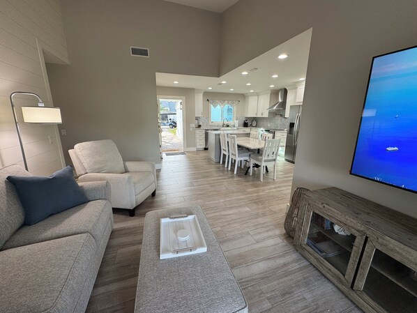A modern living room with a sofa, coffee table, and TV. It leads to a kitchen with white cabinets and a dining area.