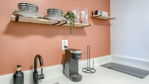 Een keuken met een wit aanrecht, een koffieapparaat, een spoelbak en een wandplank met servies en een plant.