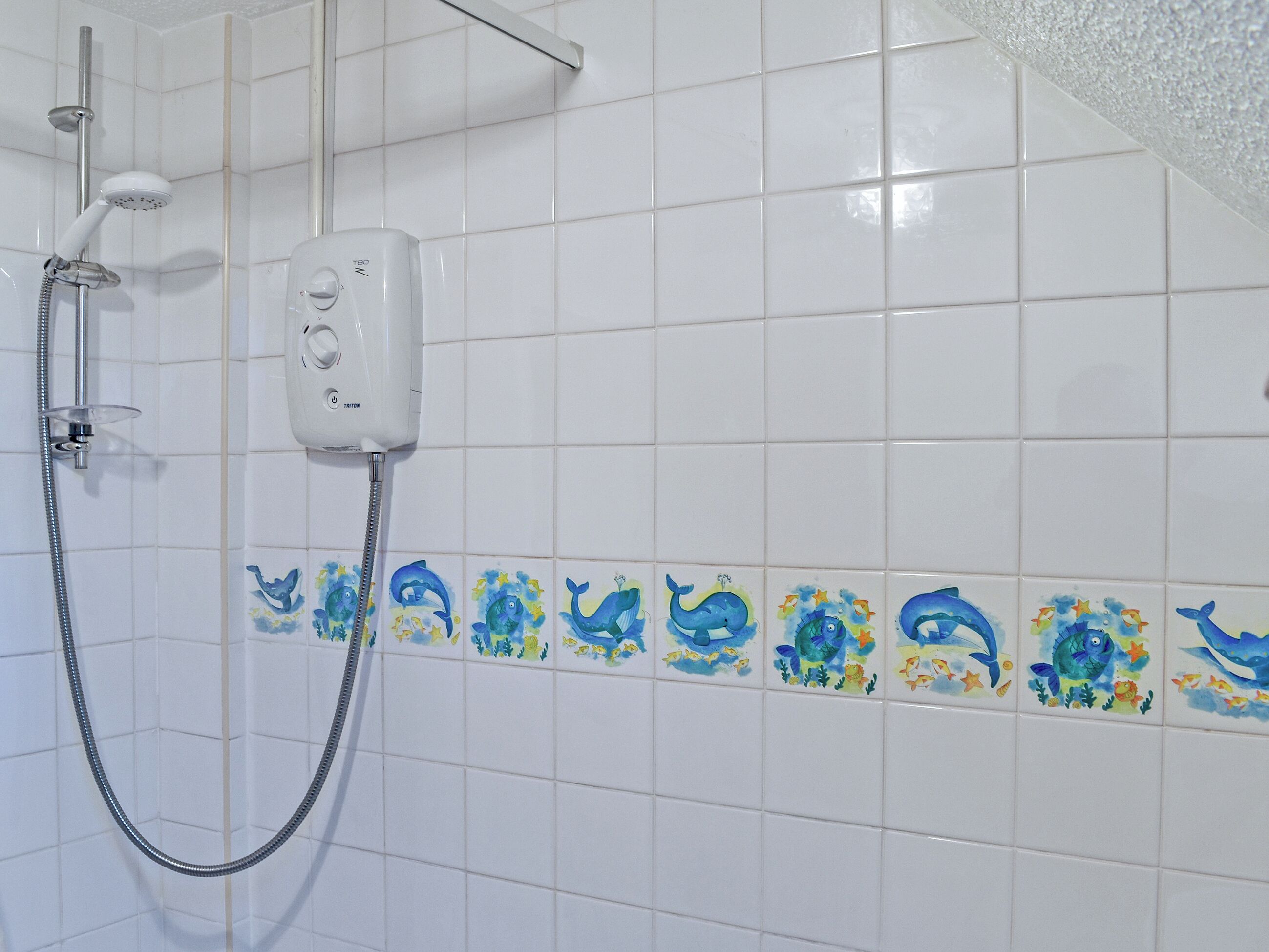 A bathroom with white tiles, a shower with a handheld hose, and a wall-mounted control panel.