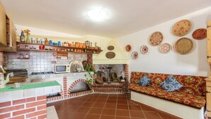 Une cuisine avec une cheminée en briques, une étagère en bois où sont rangés les ustensiles de cuisine, et un canapé rouge agrémenté de coussins à motifs.