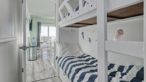 A bunk bed room with a striped bedspread, a white door, and a view into another room with a chair and a trash can. - New owner Beautiful Beach view,Indoor pool & Hot Tub (Gulf Shores)