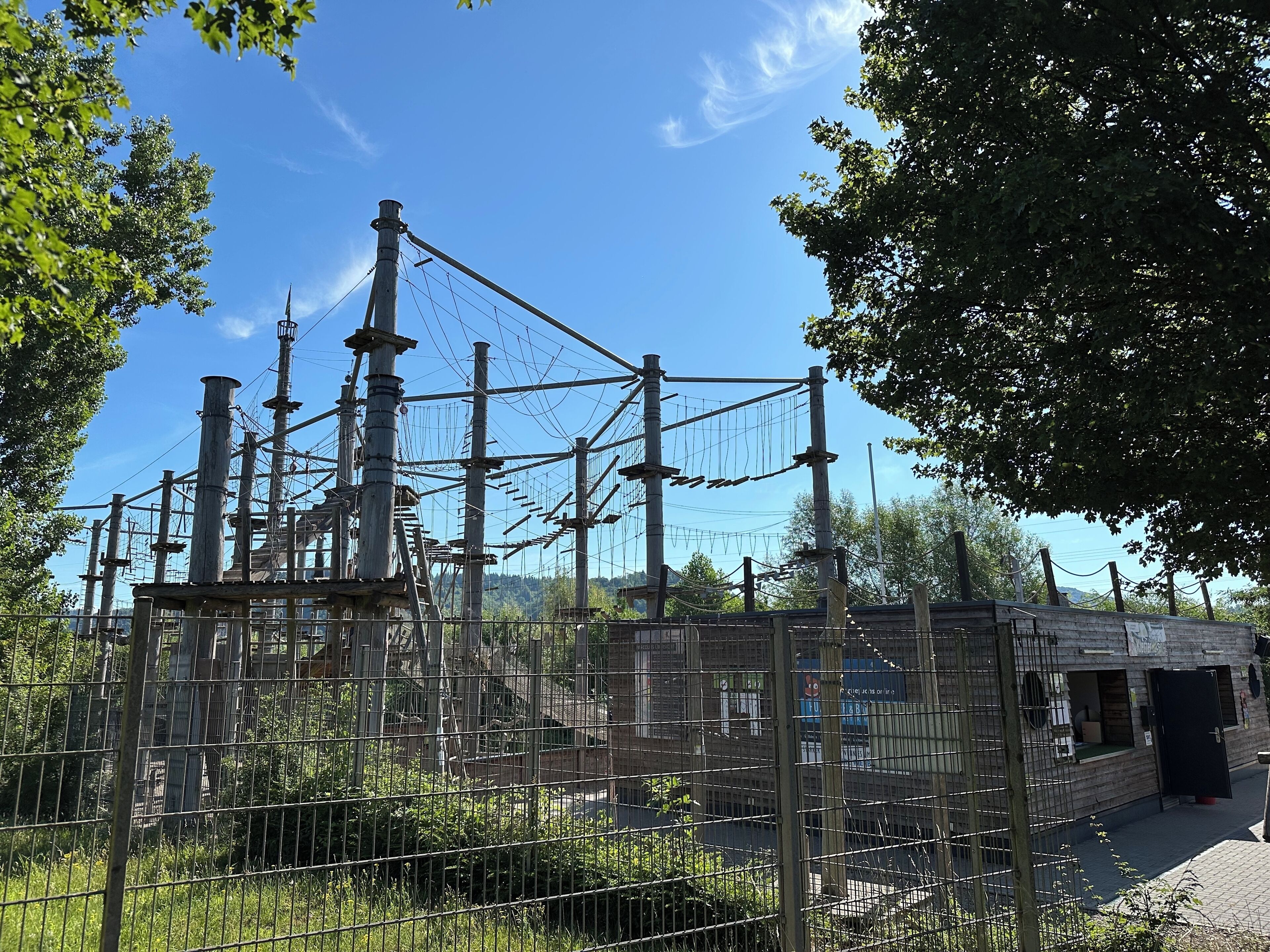 A rope bridge structure with multiple poles and cables, surrounded by trees and a building in the background.