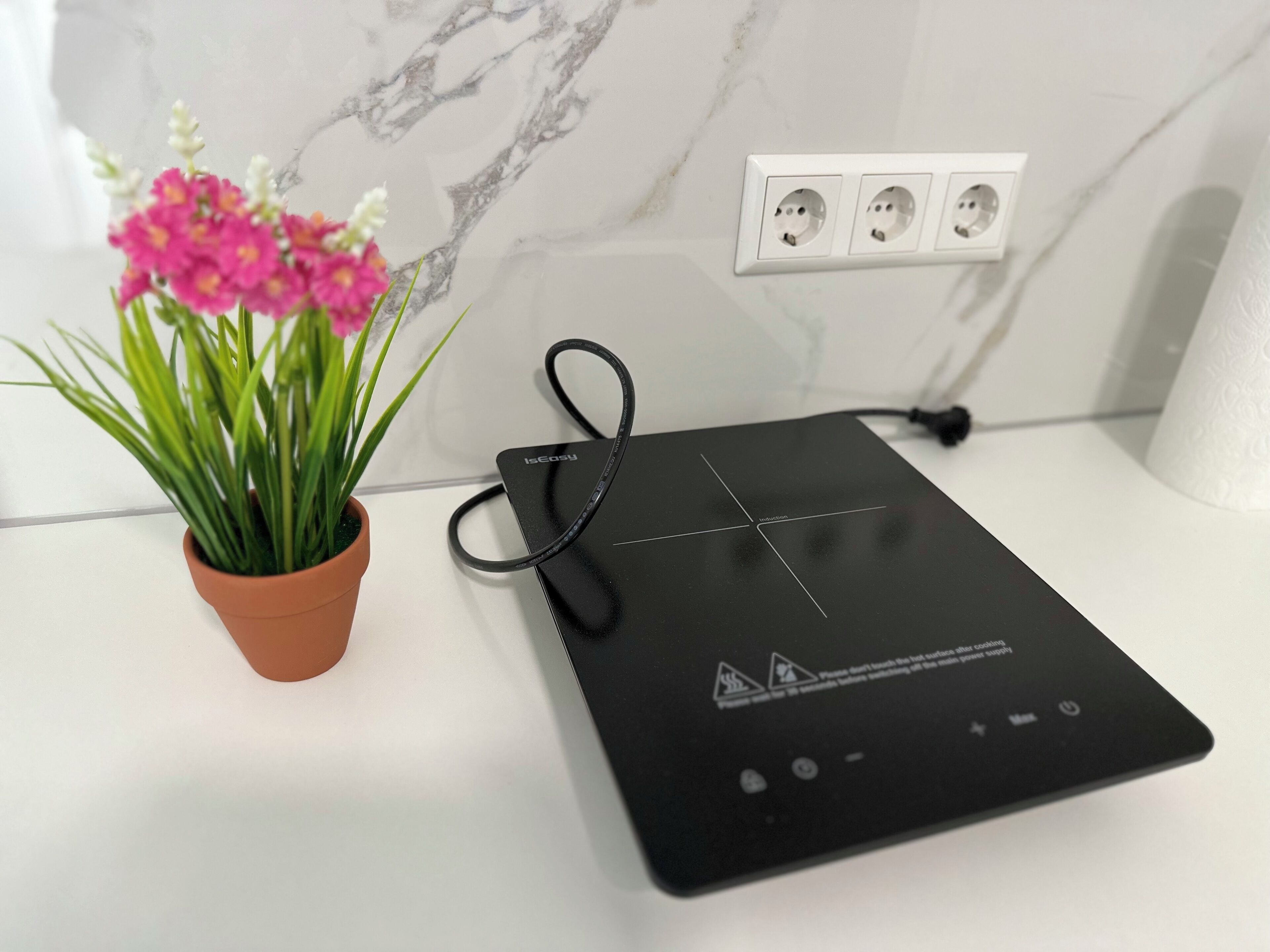 Una cocina con una placa de inducción negra, un cable de alimentación, una planta en maceta con flores rosas y un salpicadero blanco con un diseño similar al mármol.