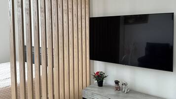 A modern living room with a large flat-screen TV mounted on the wall, a wooden slat partition, a grey console table with a potted plant, and a small dog figurine.
