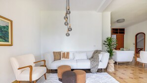 A modern living room with a white sofa, two ottomans, a white rug, and a hanging light fixture. - Spacious Villa with Pool, BBQ and Tennis Courts (Lazio)