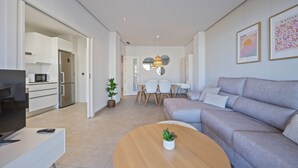 A modern living room with a grey sofa, a wooden coffee table, and a TV on a white cabinet. There is a dining area with a white table and chairs in the background.