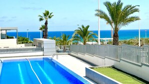 A swimming pool with clear blue water, surrounded by a white fence, palm trees, and a view of the ocean.