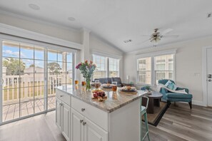 A spacious kitchen with a large island, a dining area with a blue chair, and a view of a residential neighborhood.