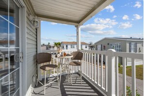 A covered porch with two wicker chairs and a small table, overlooking a row of mobile homes. - Sunny Gulf Coast Getaway-Near Beach-Shared Pool! (Fort Myers Beach)