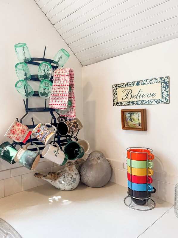 A kitchen with a variety of mugs and dishes on shelves, a decorative sign saying "Believe", and a framed picture on the wall.