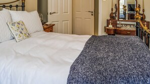 Un lit bien fait, avec du linge de lit blanc et un coussin à motifs floraux, une commode avec un miroir et des objets décoratifs, et une porte donnant sur une autre pièce.