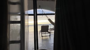 A view from inside a room with a large window overlooking a beach. There is a hammock tied to the window frame and a chair visible outside.