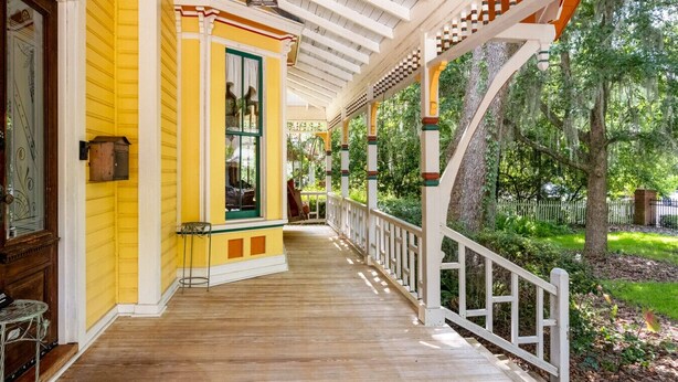 A covered porch with a wooden floor, white railings, and a yellow building with a green window. - GNV Downtown Estate- Sleeps 16 - Groups & Events (Gainesville)