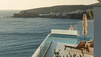 A modern poolside area with lounge chairs and an umbrella, overlooking the sea and a coastal village.