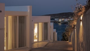 A coastal building with large sliding glass doors, a narrow pathway leading to the sea, and a view of a town across the water.
