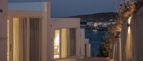 A coastal building with large sliding glass doors, a narrow pathway leading to the sea, and a view of a town across the water.
