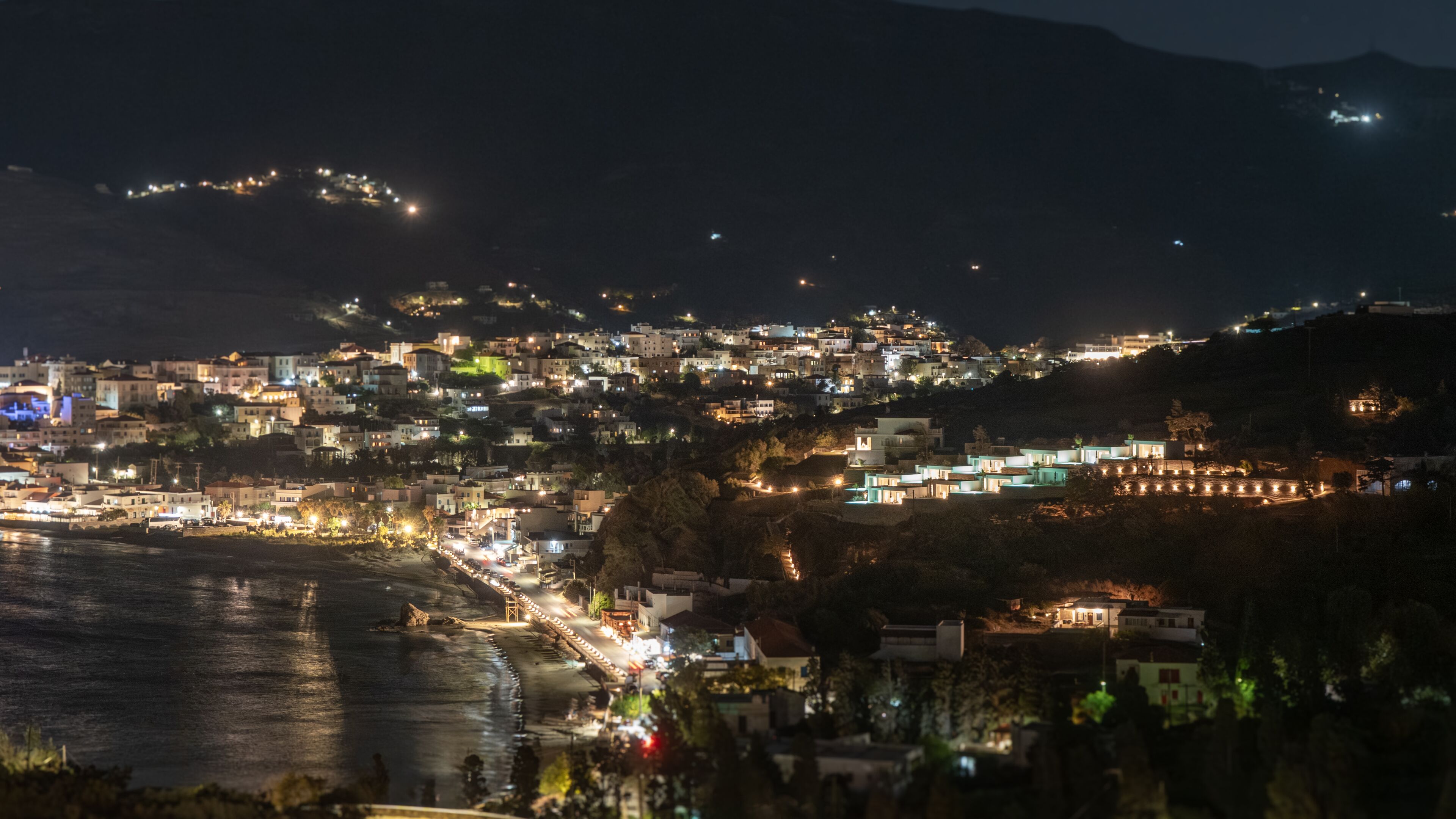Un pueblo costero de noche con edificios a lo largo del paseo marítimo, un puente y un paisaje montañoso al fondo.