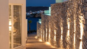 A stone-walled pathway leading to a building with a glass door, overlooking a coastal town at night.